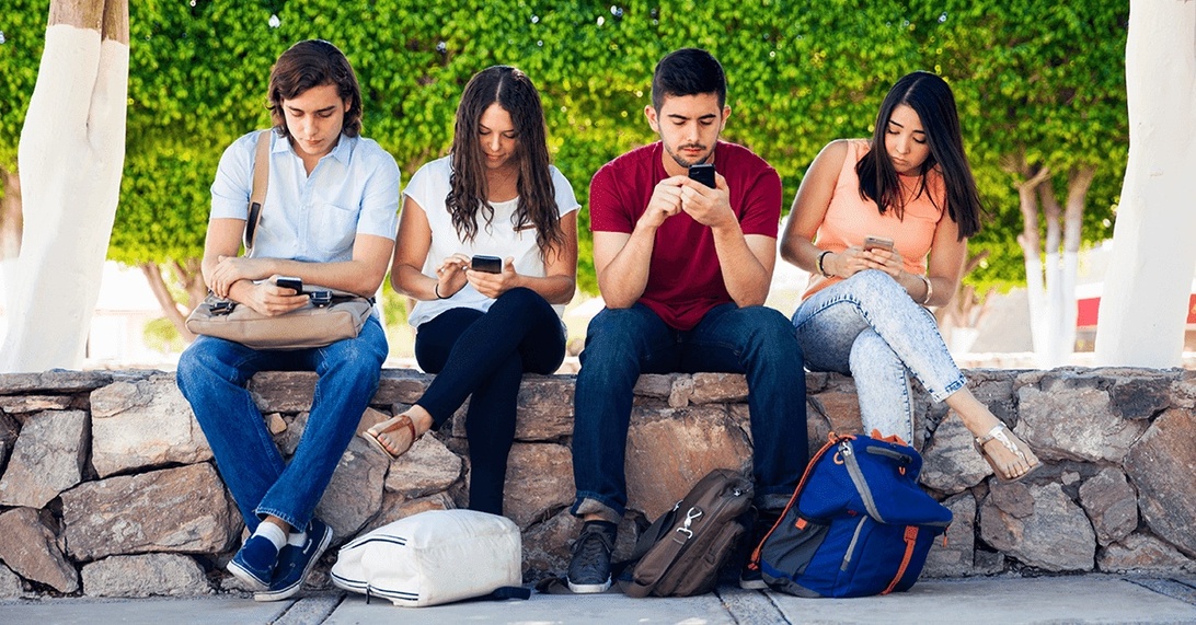 Four young adults sitting side by side on a stone wall, all looking down at their smartphones instead of engaging with each other. This powerful visual commentary shows how digital communication can replace direct human interaction, creating a scenario where people are physically together but mentally engaged in virtual spaces.